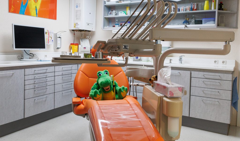Einer der Behandlungssessel der Praxis in der Farbe Orange, auf dem ein Korkodilstoffier mit Zahnbürste sitzt.